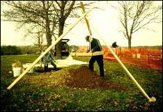 Photograph of a man leaning over a screening tool suspended by a wooden tripod, with a large pile of dirt underneath