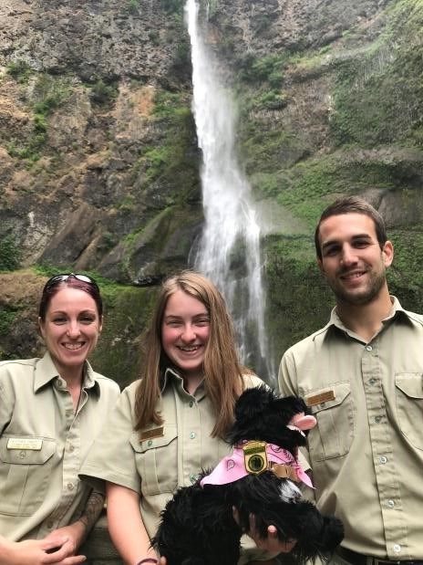 three park rangers holding a toy dog with waterfall in the background