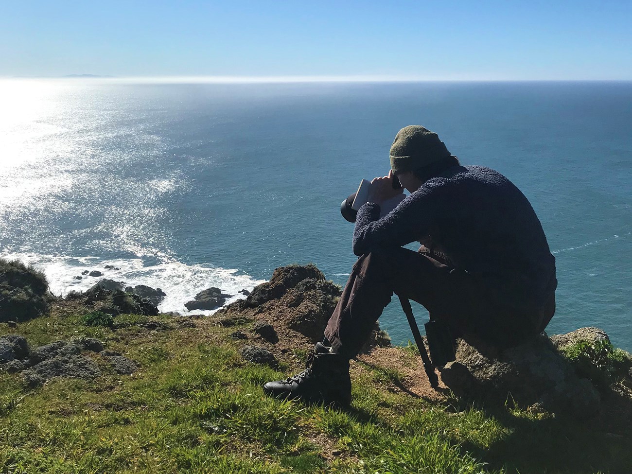 A person sits at a cliff edge while looking through a telescope.