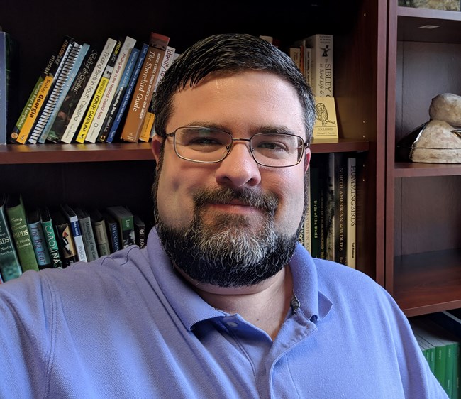 Man with a beard smiling in front of a shelf of books