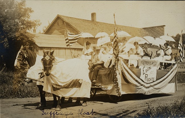 Two horses pull a float carrying about ten women in white holding parasols. Sign reads "Nebraska Next: Equal Rights."