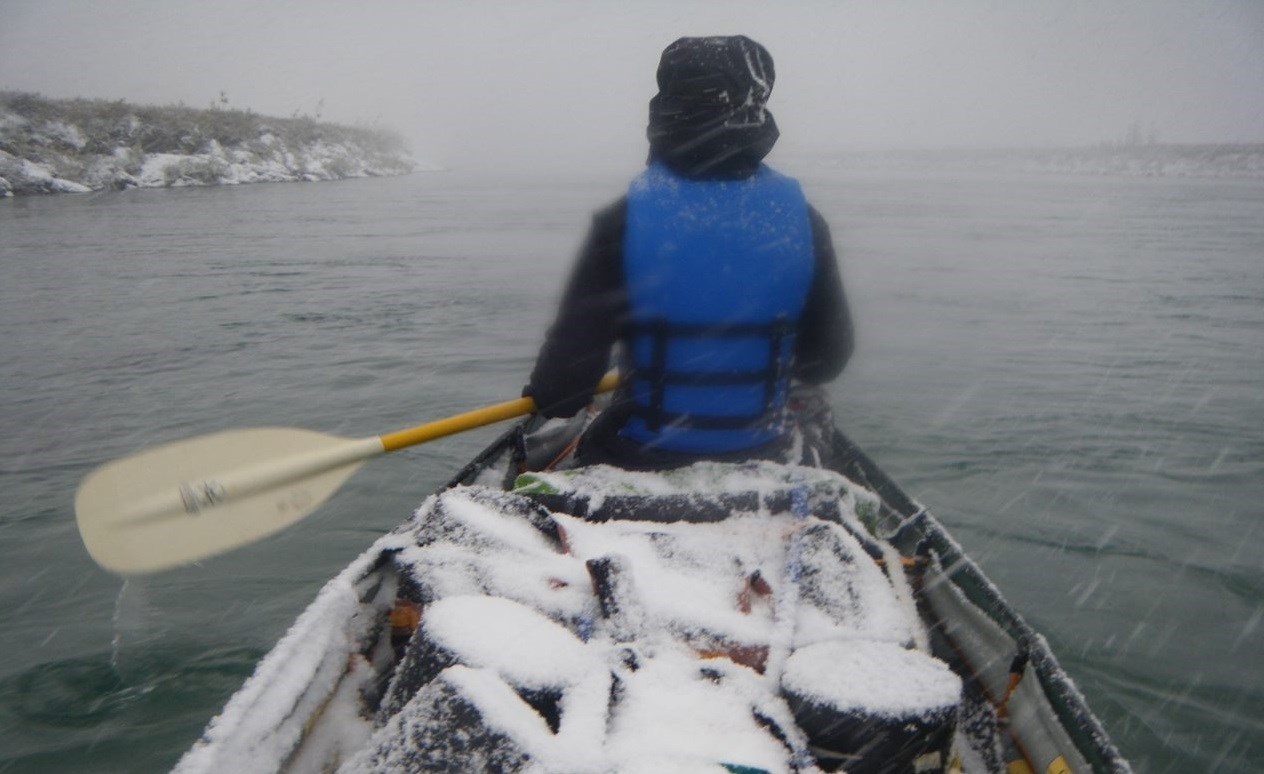 A person sits in a canoe paddling along a river forward as snow falls, creating a layer of white on all the gear packed in the boat