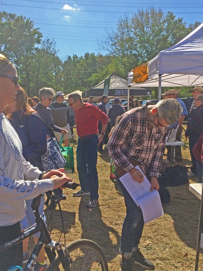 People are outside next to pop-up tents. Man on a bike, man signing a clipboard and people talking.