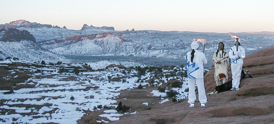 two people with Olympic torches and a Lakota flute player stand on a rock ledge