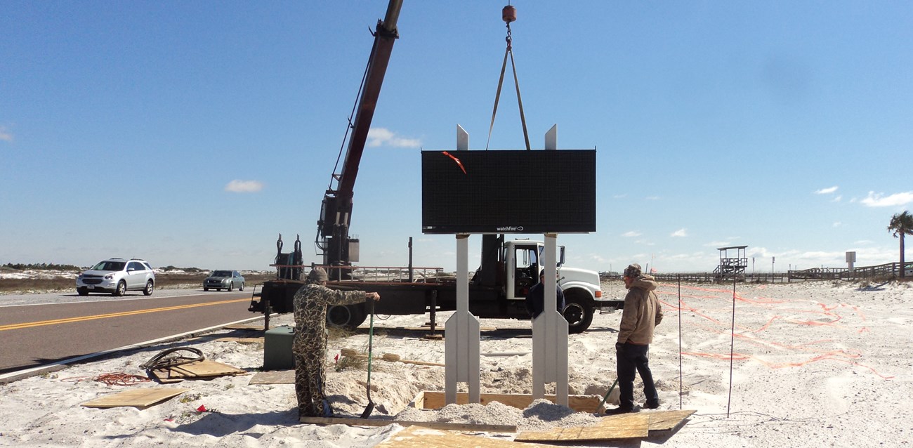 Crews install an electronic sign in sand along a road shoulder.