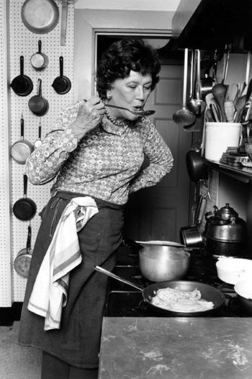 Black and white photo of Julia Child standing by her stove. She is tasting something with a spoon. A skillet on her stove has something cooking in it. She has a towel tucked into her apron strings. She is surrounded by cooking utensils.
