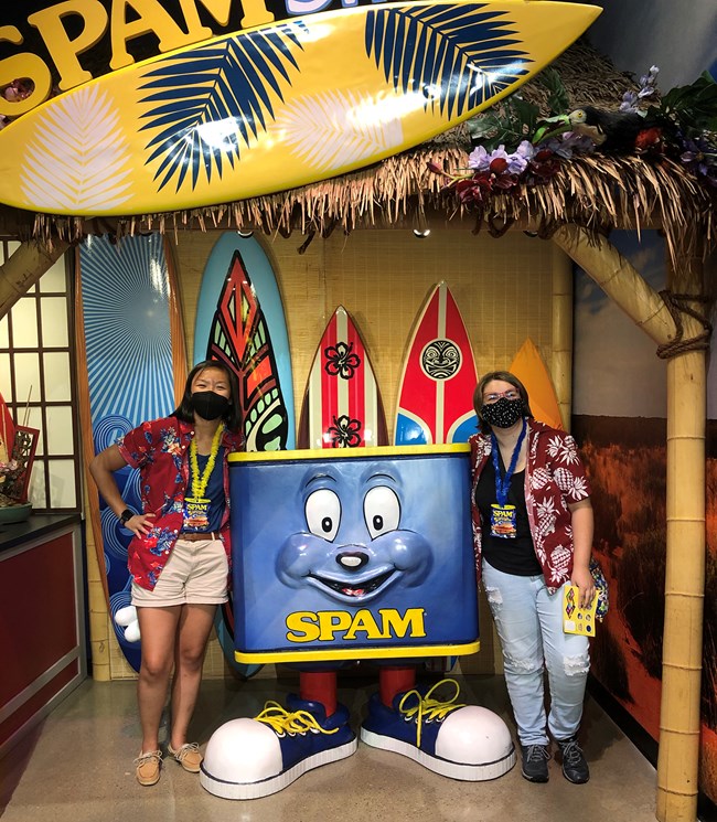 Two people in Hawaiian shirts pose beside a smiling character (a can of Spam with a face and legs, wearing sneakers). Behind them are surfboards.