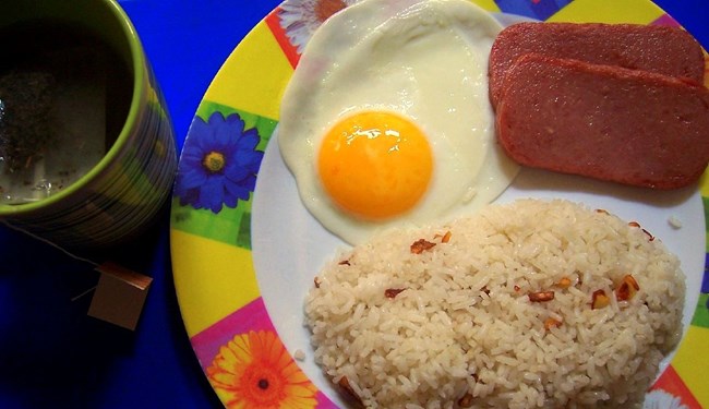 A color photo showing spamsilog served on a colorful plate. A mug of tea is to the left of the plate.