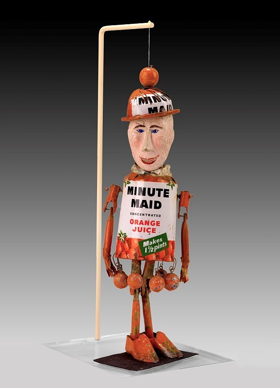 Color photo. Can (black, orange, and green) reads “Minute Maid Concentrated Orange Juice. Makes 1-1/2 Pints.” Arms, legs, and feet are colored orange. The hat, also from a can, reads “Minute Maid.” Face made of painted wood.