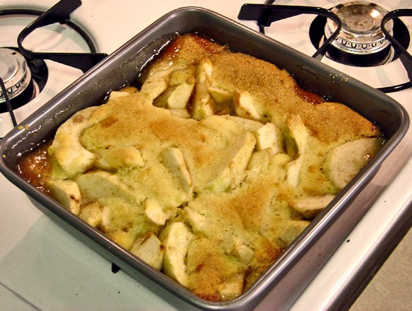 A square baking pan sitting on top of a white gas stove. In the pan is apple cobbler – slices of apples covered with a thin crust.