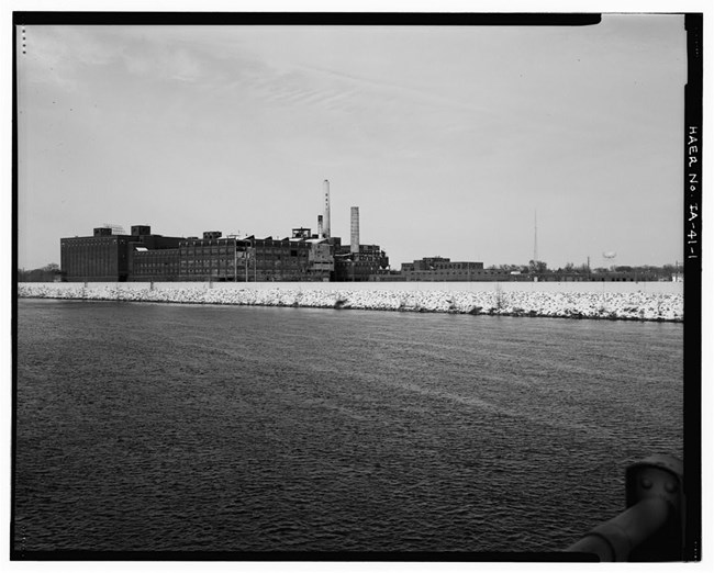 Black and white photograph of ocean in the foreground and a large building in the background.