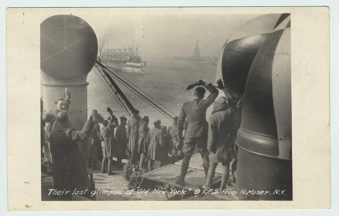 A boat full of soldiers in WWI uniforms wave to the Statue of Liberty as they pass