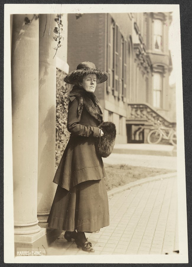 Informal portrait, full-length, Sallie Hovey of New Hampshire, facing right with head turned toward camera, standing outside by column of what is likely National Woman's Party headquarters, wearing two-piece suit with long skirt, fur stole, wide-brimmed h