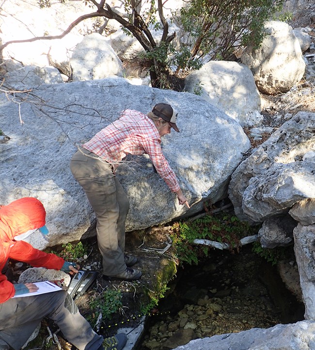 Two people with water monitoring equipment and a clipboard standing next to a pool of water surrounded by large boulders.