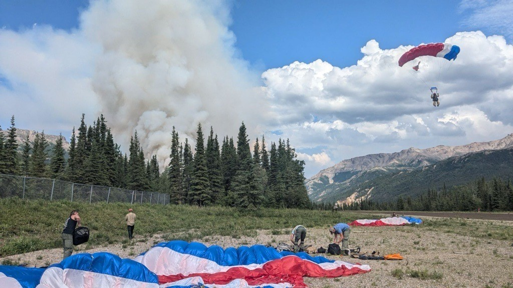 A smokejumper descends to the ground via parachute, while four others prepare their gear on the ground. A plume of smoke rises out of the forest behind them.