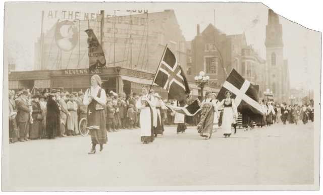 Women in Scandinavian dress holding flags marching down street with crowds on sidewalk