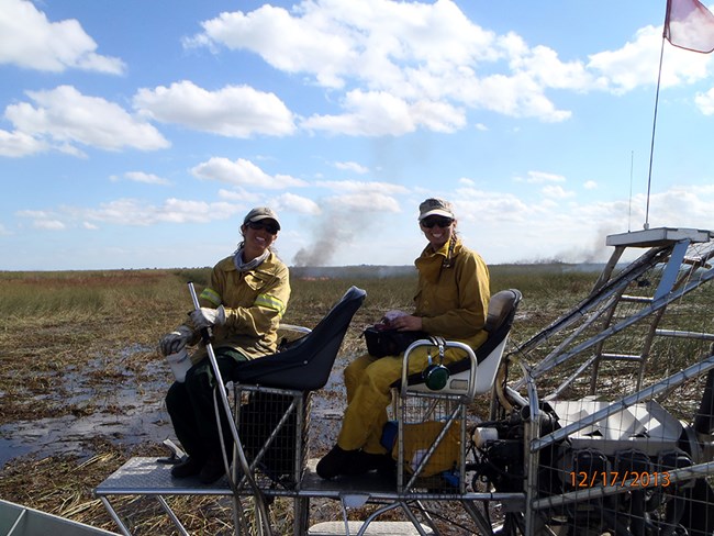 Two women sit in a vehicle in a wetland with a fire in the background.