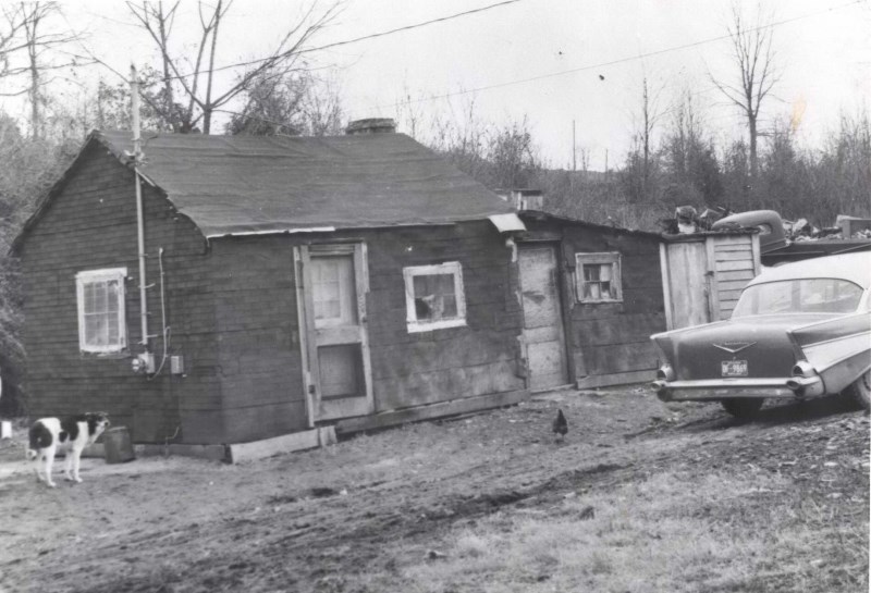 Black & white photo of a typical Tobytown house. Small house with an attached garage. The house is in poor condition with warping wood and missing window panes with bare trees behind and an equally bare yard in front. A dog, chicken, & car are in the yard