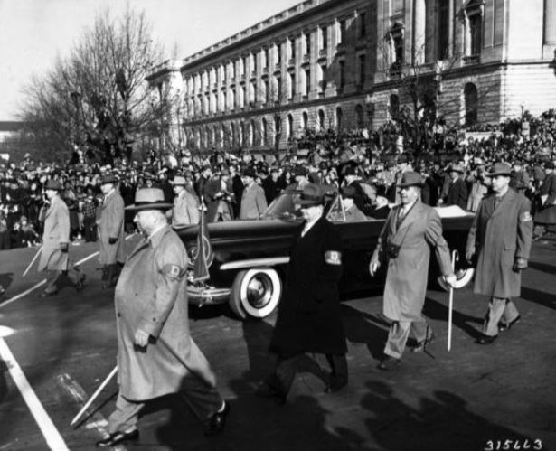 Harry Truman rides in a convertible car on Pennsylvania Avenue, flanked by men in trench coats