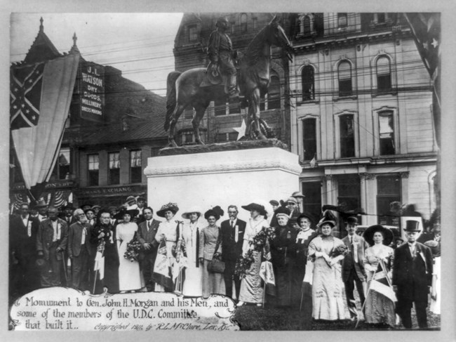 Men and women stand in front of General Morgan's monument. Dressed in fancy clothes with Confederate memorabilia.