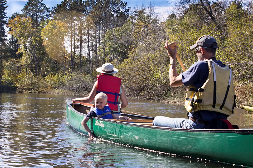 Family in green canoe on tree lined river, child looking back at camera