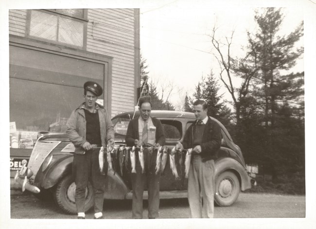 Black and white photo of three men holding a stick with a dozen fish hanging down vertically, in front of an old-fashioned car and building.