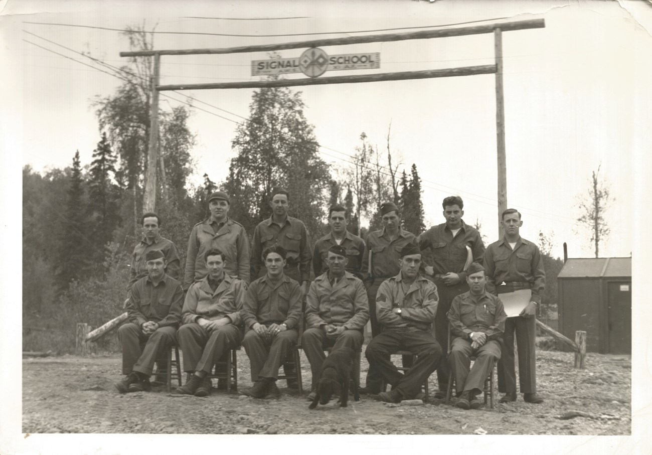 Black and white photo of uniformed men, six seated in front of seven standing behind them, below a wooden gateway that says "Signal School."