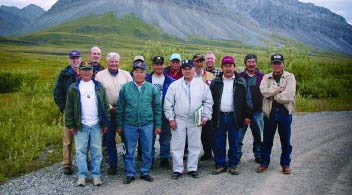 A group of 14 men stand on a gravel road in front of a mountain.