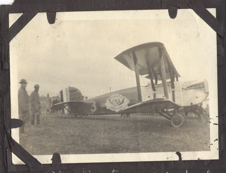 Black and white photo of biplane aircraft. A group of people stand behind the aircraft.