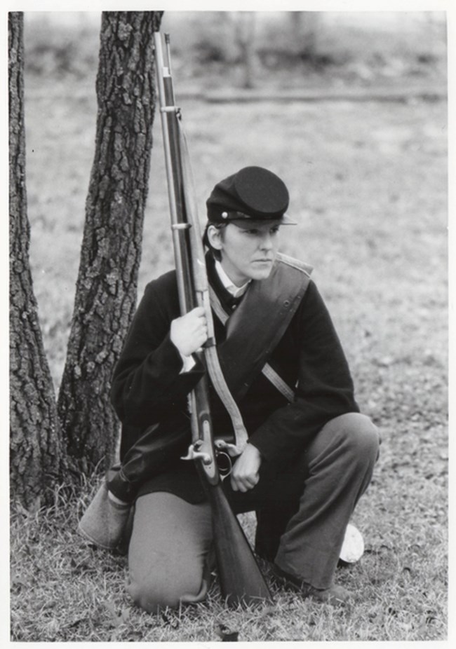 Black and white photo of Lauren Cook posing in Civil War uniform. She is kneeling in front of a tree, holding a period rife, looking off into the distance. Expansive battleground behind her