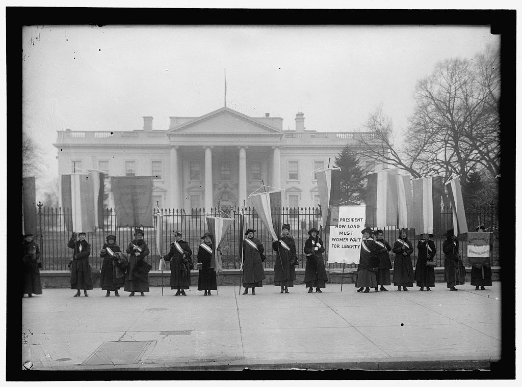 Suffragists picketing outside of the White House. Some women hold flags in suffrage colors and one woman holds a banner reading, "Mr. President how long must women wait for liberty."