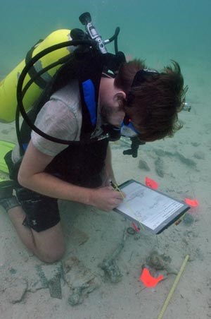 [photo] SCUBA diver writing on tablet at underwater archeological site.