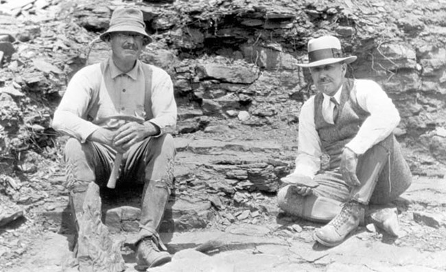 A black and white image of two men seated on rocks. A slab with fossil plant fronds is propped in front of the right leg of the man on the left.