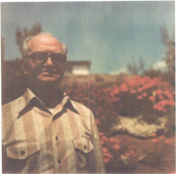 Color photo of an older man posing in front of brightly colored flowers.