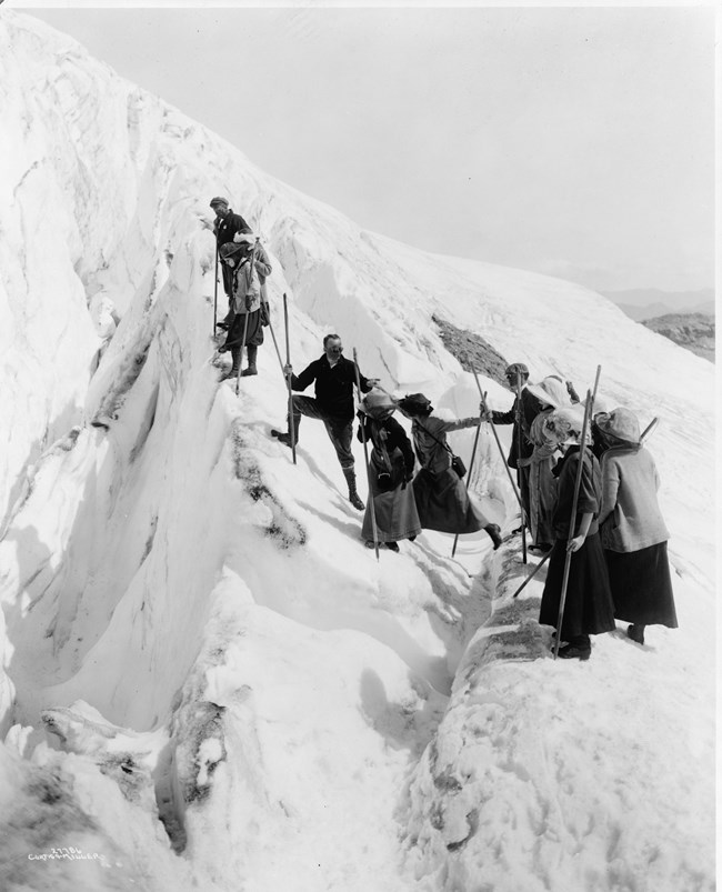 A group of climbers in historic clothing stand at the edge of a snowy precipice on a glacier. Some look over the edge while others move closer to the edge.