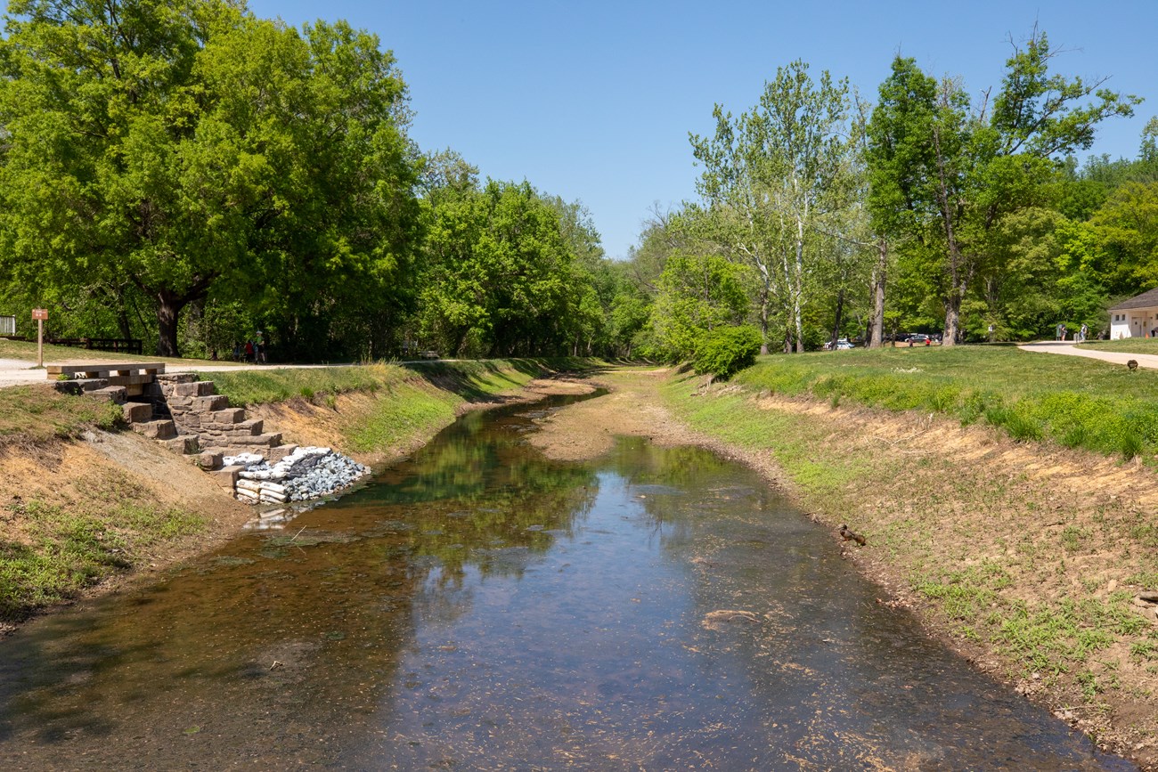 A partially full of water canal prism that extends away from view. The towpath is located to the left of the canal with a paved sidewalk on the right.