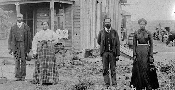Two Black men and two black women stand in front of a frame building. Photo is black and white.