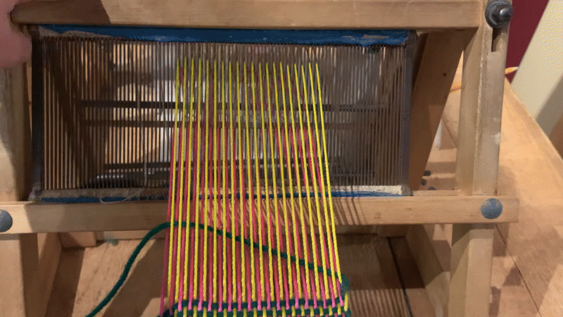 gif of a metal comb like bar with vertical yellow and pink threads running through it is pulled forward, pushing a horizontal green yarn forward to join woven material