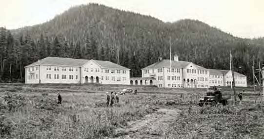 Black and white photo of large buildings in front of a forested hill