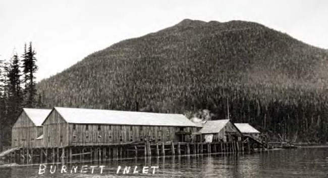 Black and white photo of long wood buildings on piers in front of trees