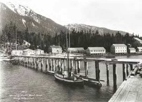 Black and white photo of a pier with buildings along the shore and mountains behind