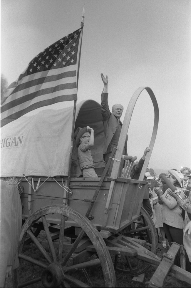 A man in a modern suit stands in a covered wagon and waves his hand.