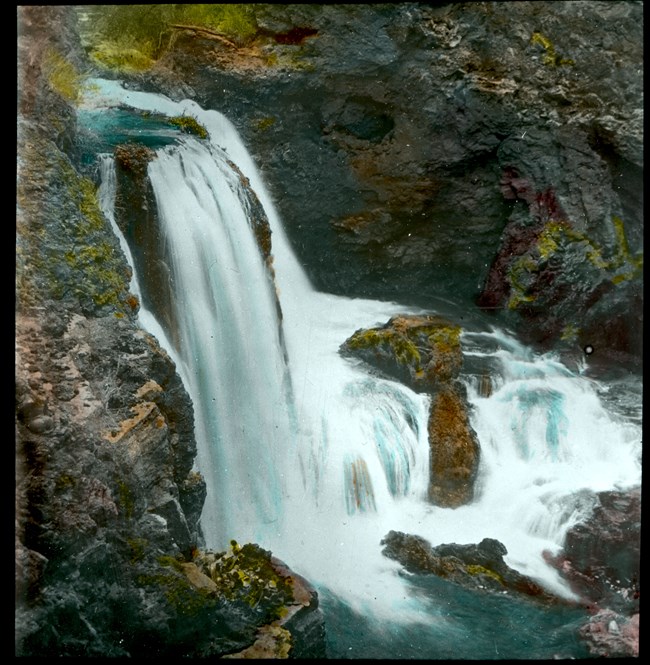 A waterfall pours over dark rocks into a clear blue pool.