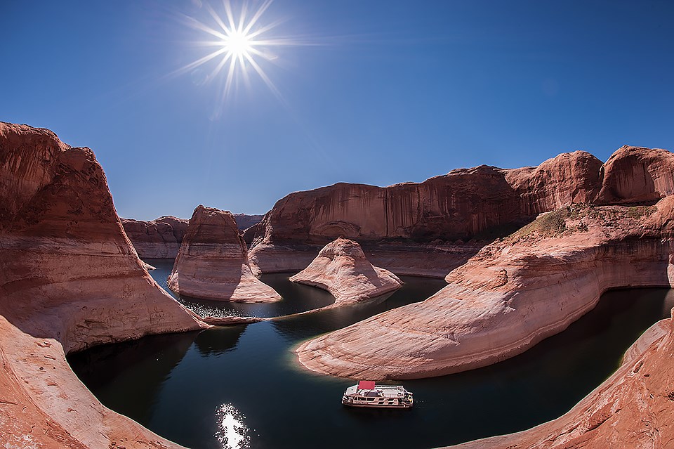 A winding river through steep red rocky cliffs with a boat in the water and a shining sun in the sky