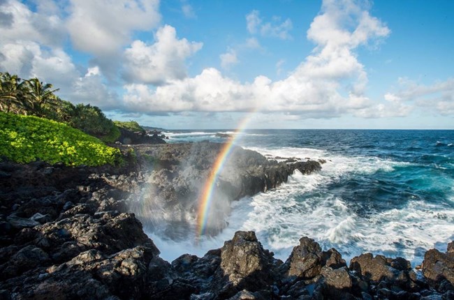 A rainbow forms in the mist of the ocean crashing on a rocky shore