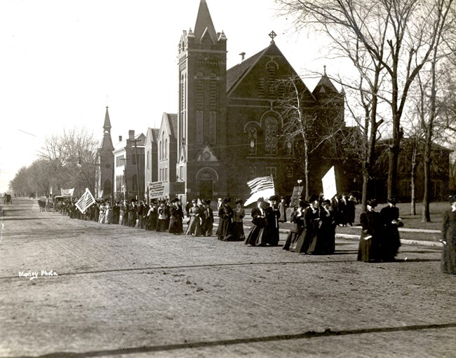 Woman suffrage procession passing by church. Women carrying flags and banners
