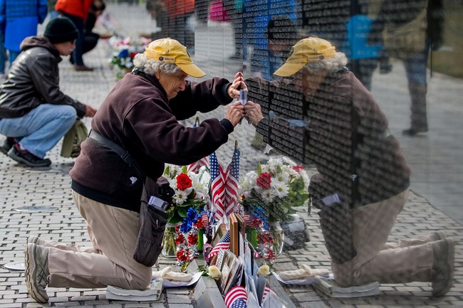 A woman in volunteer uniform kneels near a bouquet of flowers at the Vietnam Wall to get a rubbing of a name from the wall.