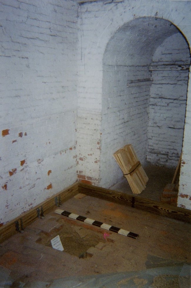 Room with whitewashed brick walls and brick floor. A group of bricks is removed at center with a hole dug out, next to a black and white scale bar.