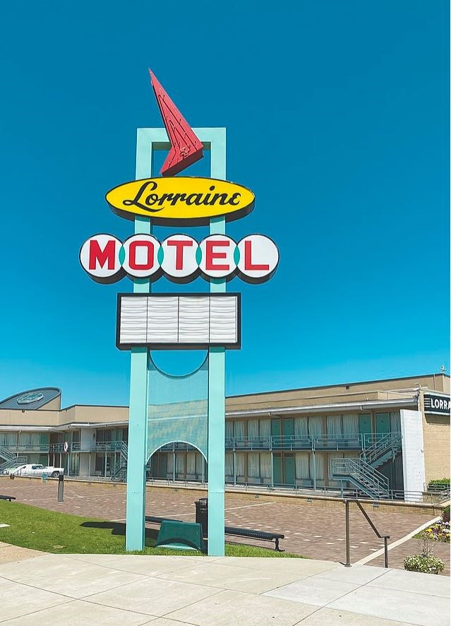 sidewalk view of the two story motel with several doors to the rooms. A blue, red and yellow sign is visible from the street and has the motel's name on it.