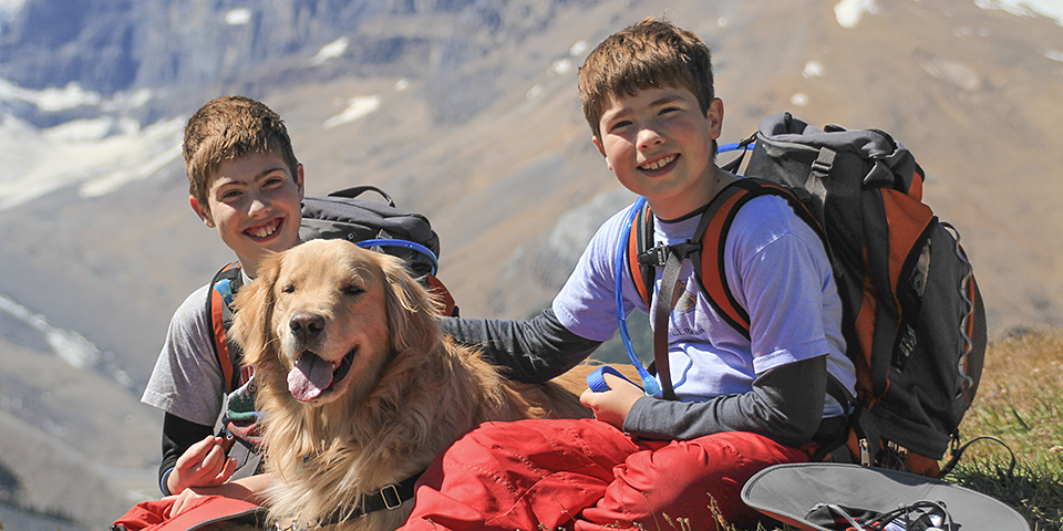 Two kids wearing backpacks sit with a leashed golden retriever
